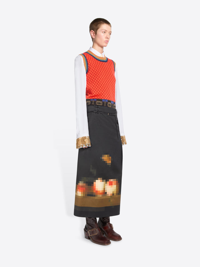 A model stands against a light gray background wearing a long black skirt with a pixelated multicolor graphic near the hem, a red patterned sleeveless top, a white long-sleeve shirt, and brown ankle boots.