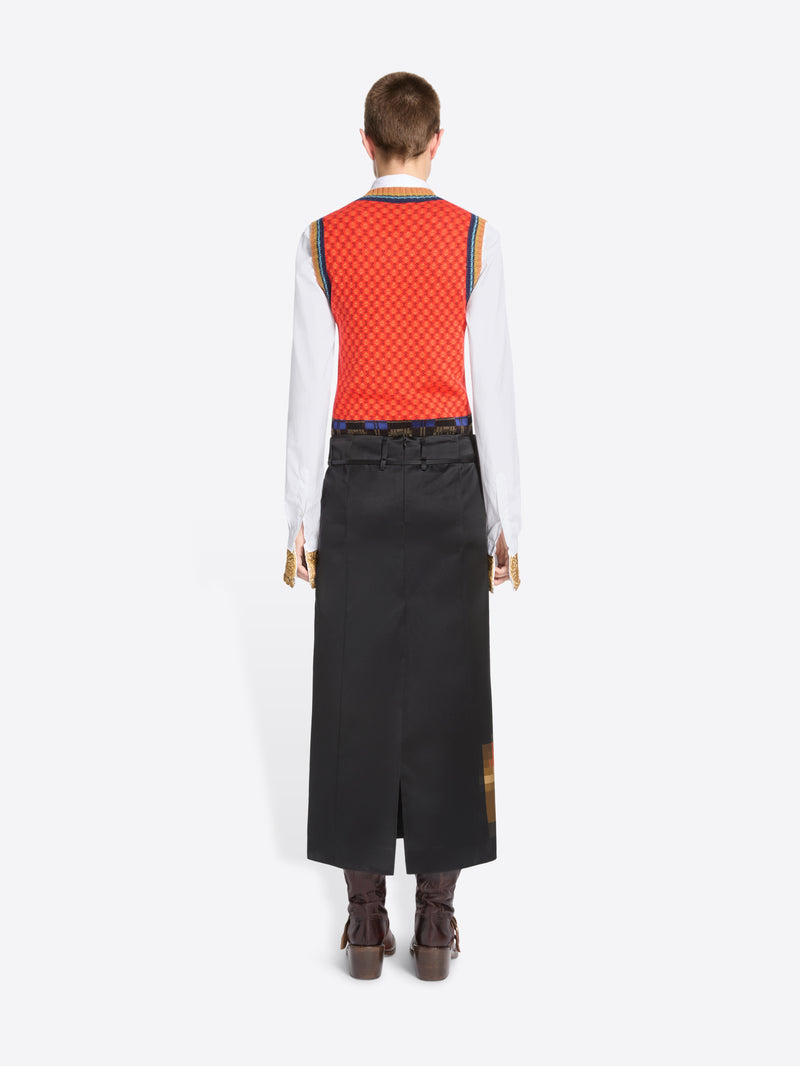 Back view of a model standing against a light gray background wearing a long black skirt with a pixelated multicolor graphic near the hem, a red patterned sleeveless top, a white long-sleeve shirt, and brown ankle boots. 