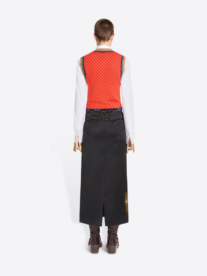 Back view of a model standing against a light gray background wearing a long black skirt with a pixelated multicolor graphic near the hem, a red patterned sleeveless top, a white long-sleeve shirt, and brown ankle boots. 