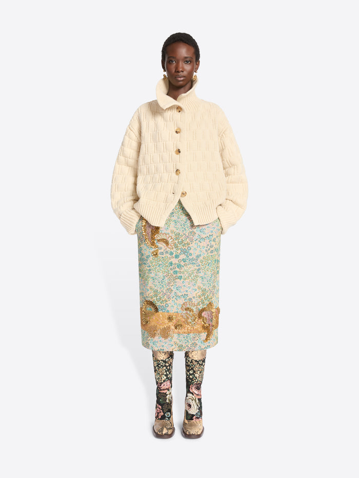 A model stands against a light gray background wearing a cream-colored textured knit cardigan over a light blue and green patterned skirt, paired with floral-patterned tights and snakeskin heels.