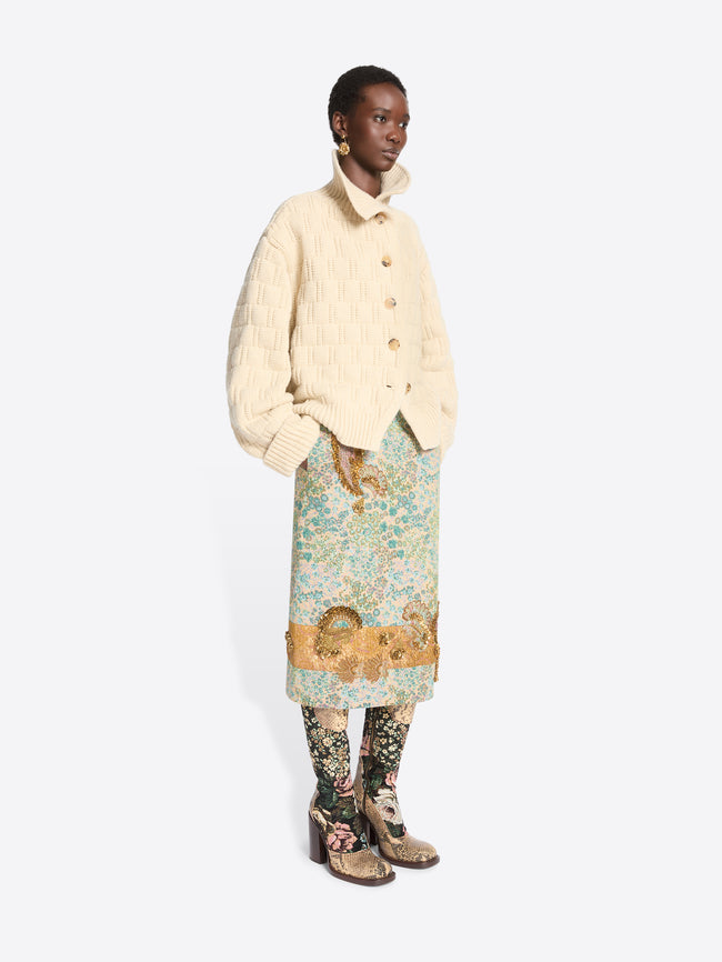 A model stands against a light gray background wearing a cream-colored textured knit cardigan over a light blue and green patterned skirt, paired with floral-patterned tights and snakeskin heels.