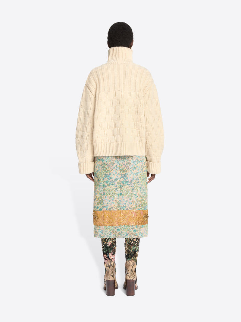 Back view of a model standing against a light gray background wearing a cream-colored textured knit cardigan over a light blue and green patterned skirt, paired with floral-patterned tights and snakeskin heels.