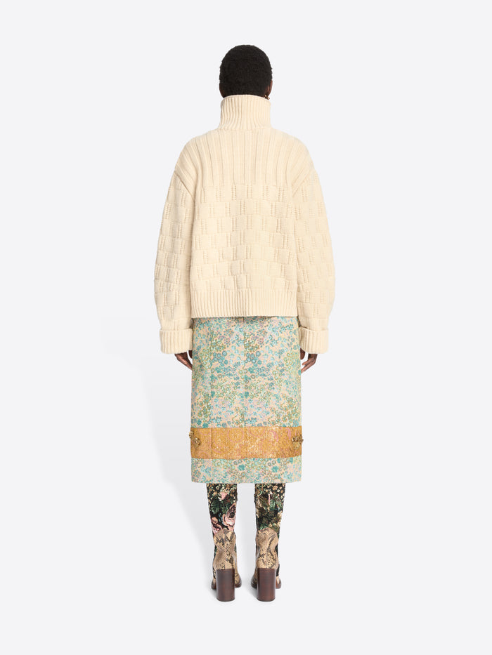 Back view of a model standing against a light gray background wearing a cream-colored textured knit cardigan over a light blue and green patterned skirt, paired with floral-patterned tights and snakeskin heels.