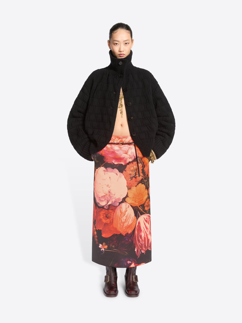 A model stands against a light gray background wearing a black cardigan and a long black skirt with a large pink, orange, and red floral print, paired with dark brown ankle boots.