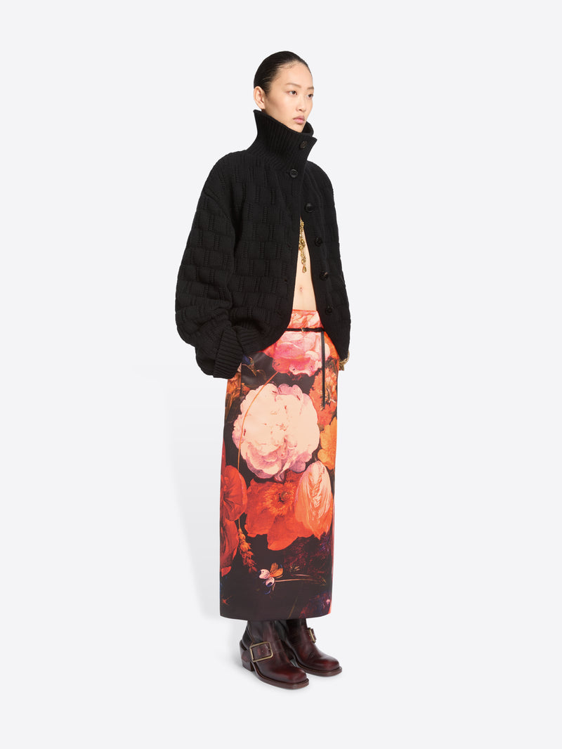A model stands against a light gray background wearing a black cardigan and a long black skirt with a large pink, orange, and red floral print, paired with dark brown ankle boots.