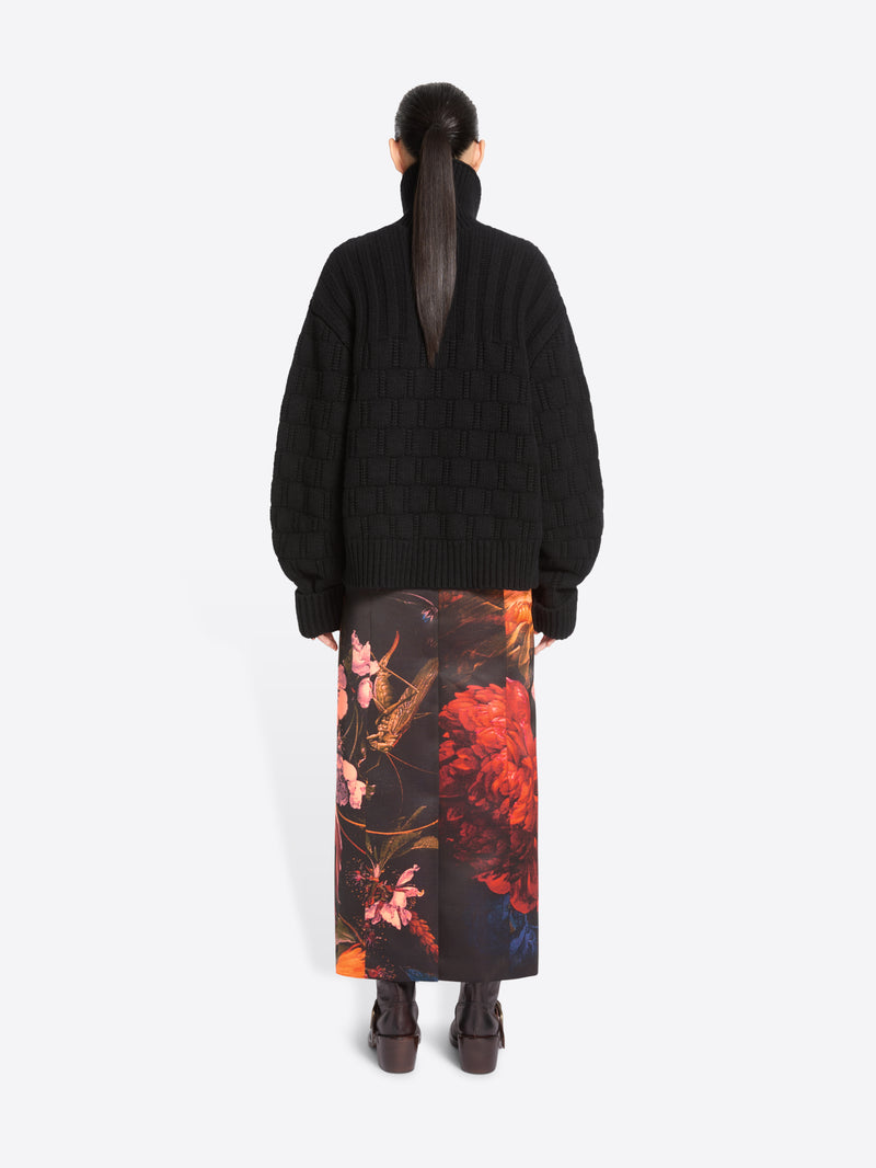 Back view of a model standing against a light gray studio background wearing a black cardigan and a long black skirt with a large pink, orange, and red floral print, paired with dark brown ankle boots.