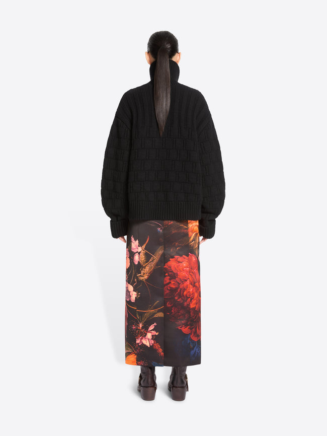 Back view of a model standing against a light gray studio background wearing a black cardigan and a long black skirt with a large pink, orange, and red floral print, paired with dark brown ankle boots.