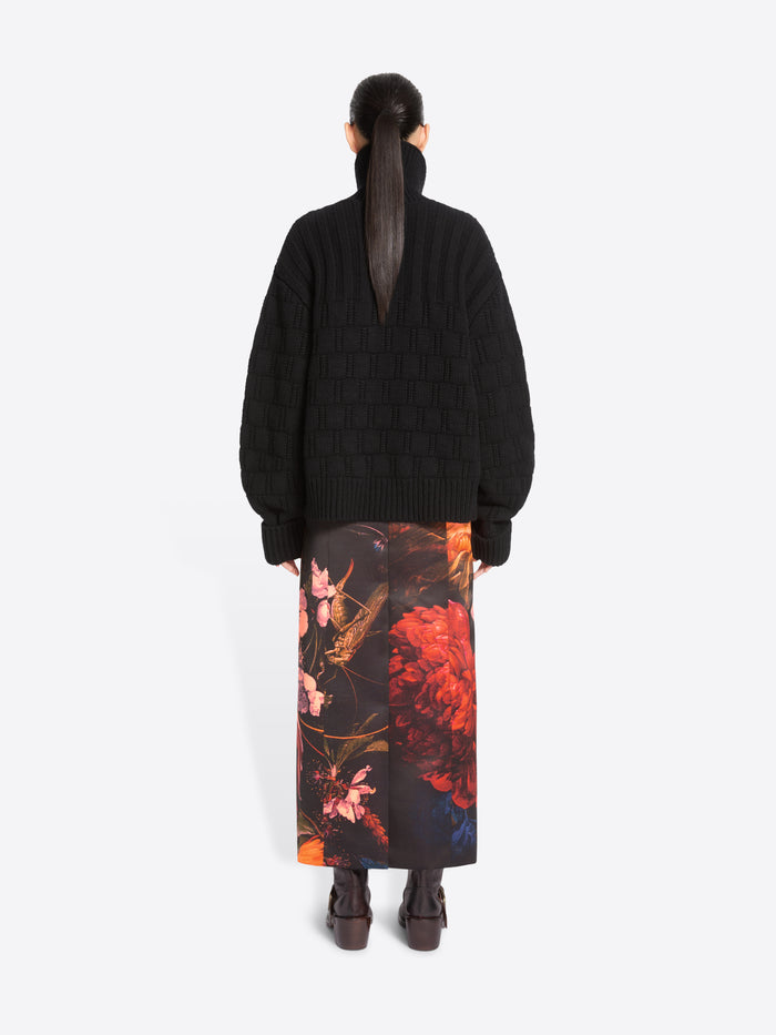 Back view of a model standing against a light gray studio background wearing a black cardigan and a long black skirt with a large pink, orange, and red floral print, paired with dark brown ankle boots.