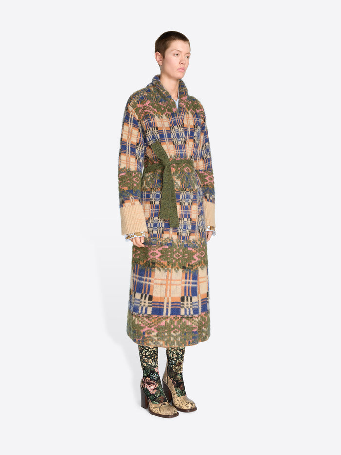 A model stands against a light gray background wearing a long patterned coat with a matching belt, paired with patterned tights and snakeskin heels.