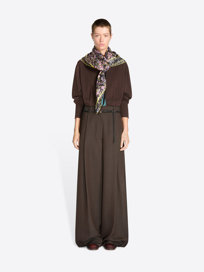 A model stands against a light gray background wearing a dark brown long-sleeve sweater, matching wide-leg trousers, a thin black belt, and a patterned scarf.