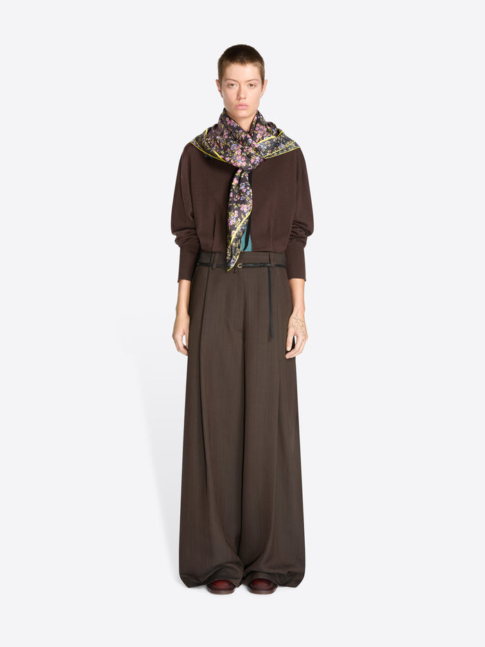 A model stands against a light gray background wearing a dark brown long-sleeve sweater, matching wide-leg trousers, a thin black belt, and a patterned scarf.