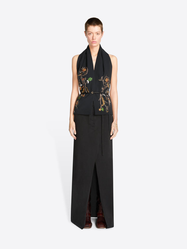 A model wearing a black sleeveless wrap-style top with ornate gold, green, and brown floral embroidery on the front, paired with a long black skirt featuring a front slit against a light gray background.