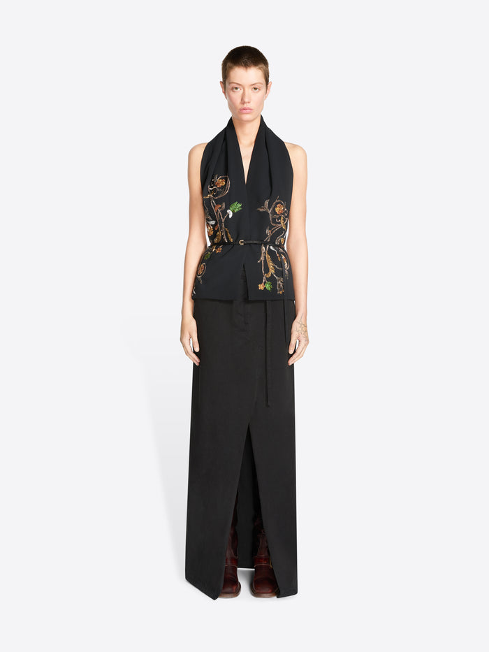 A model wearing a black sleeveless wrap-style top with ornate gold, green, and brown floral embroidery on the front, paired with a long black skirt featuring a front slit against a light gray background.