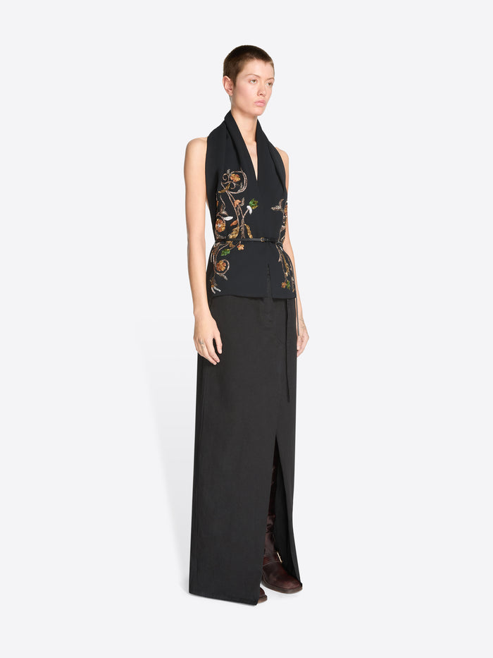 A model wearing a black sleeveless wrap-style top with ornate gold, green, and brown floral embroidery on the front, paired with a long black skirt featuring a front slit against a light gray background.
