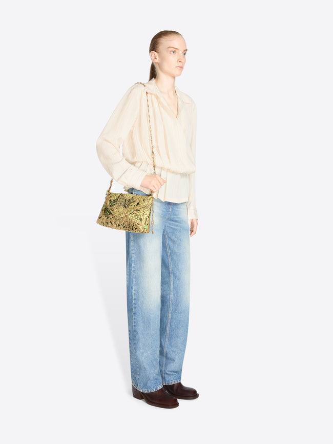 Full-length model wearing a cream striped blouse and light-wash jeans, carrying a small black-and-gold floral jacquard bag with green accents and chain handles.