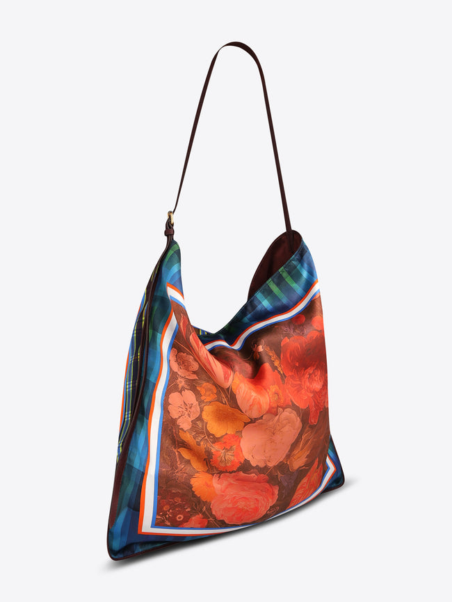 Triangular shoulder bag with a long dark brown strap, blue-green plaid panels, and a red-orange floral center framed with striped trim on a light gray background.