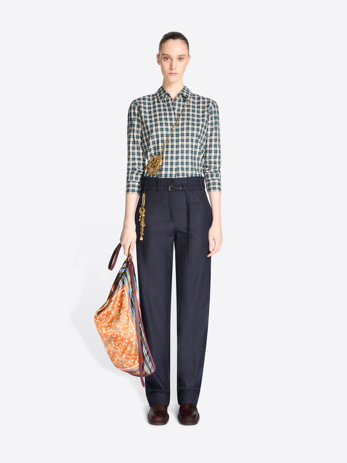 Full-length model wearing a blue checked shirt and navy trousers, carrying an orange floral triangular shoulder bag with plaid panels.