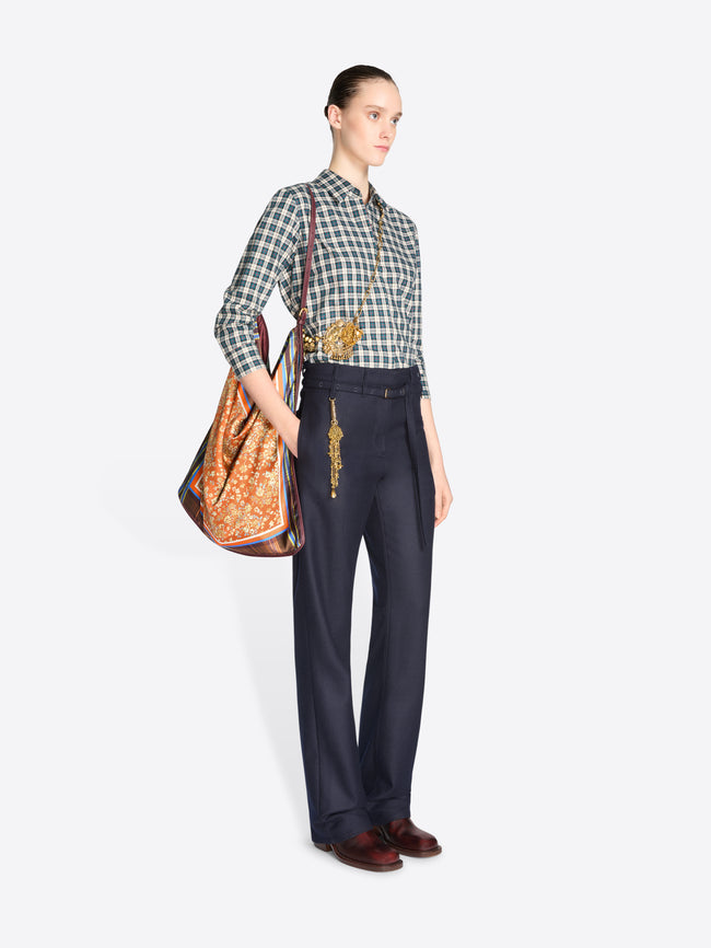 Full-length model wearing a blue checked shirt and navy trousers, carrying an orange floral triangular shoulder bag with plaid panels.
