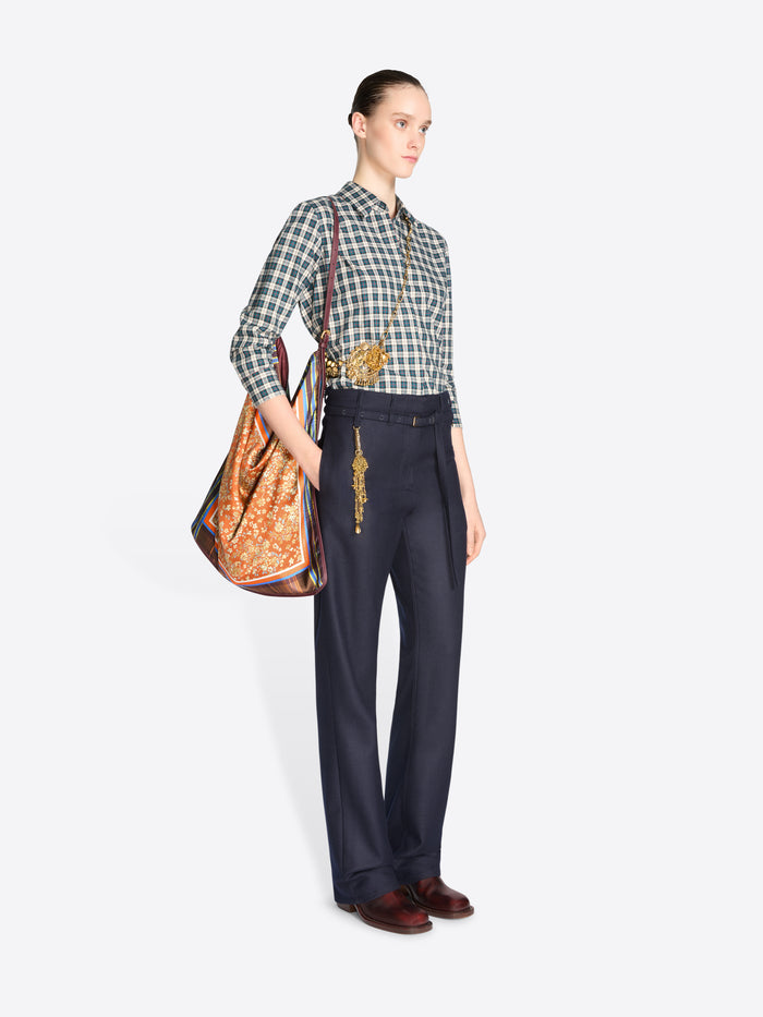 Full-length model wearing a blue checked shirt and navy trousers, carrying an orange floral triangular shoulder bag with plaid panels.