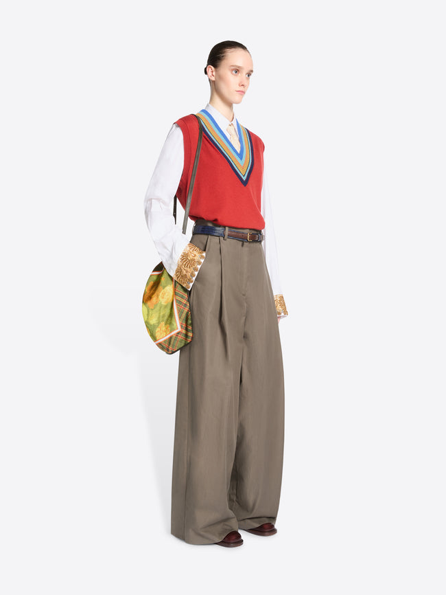 Full-length model wearing a red V-neck sweater vest, white shirt, and wide-leg taupe trousers, carrying a green plaid triangular shoulder bag with a yellow floral panel.