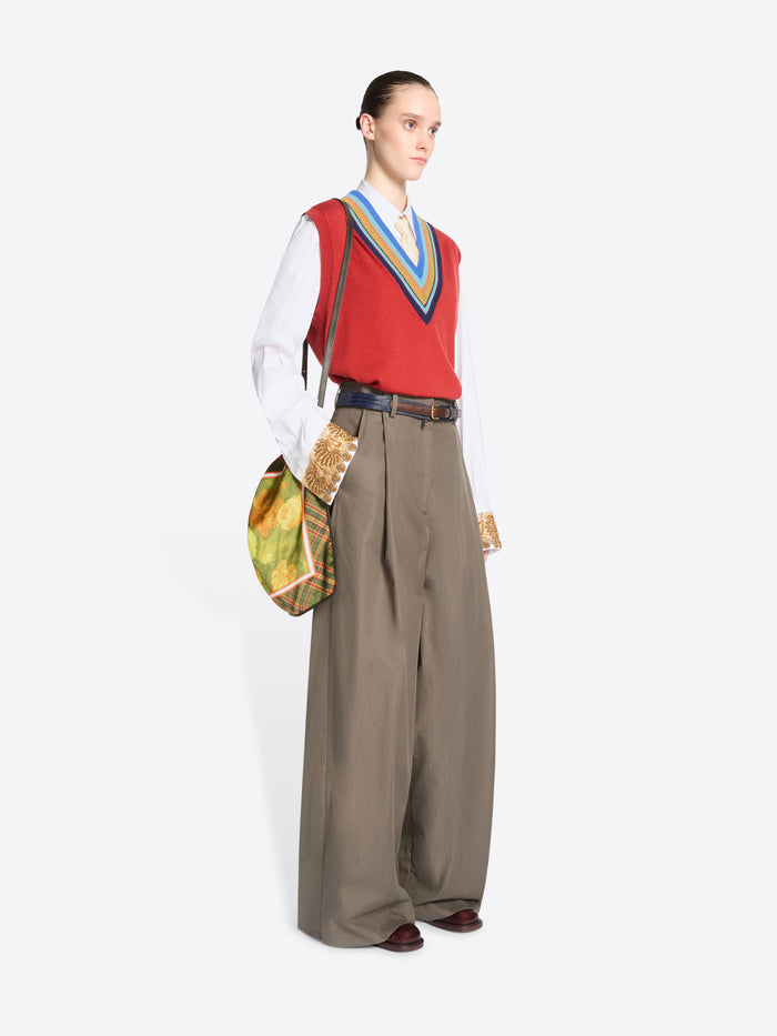 Full-length model wearing a red V-neck sweater vest, white shirt, and wide-leg taupe trousers, carrying a green plaid triangular shoulder bag with a yellow floral panel.