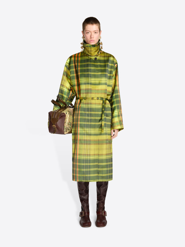 Full-length model wearing a green plaid belted coat, long brass earrings, and dark brown knee-high boots, carrying a floral jacquard duffle bag with tan snakeskin-effect panels.