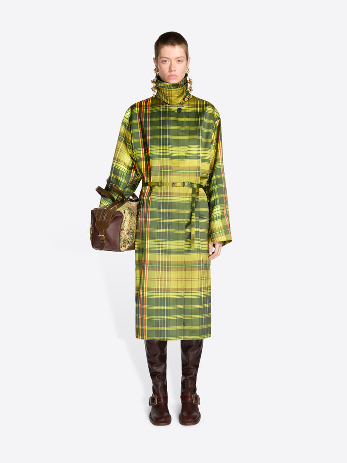 Full-length model wearing a green plaid belted coat, long brass earrings, and dark brown knee-high boots, carrying a floral jacquard duffle bag with tan snakeskin-effect panels.
