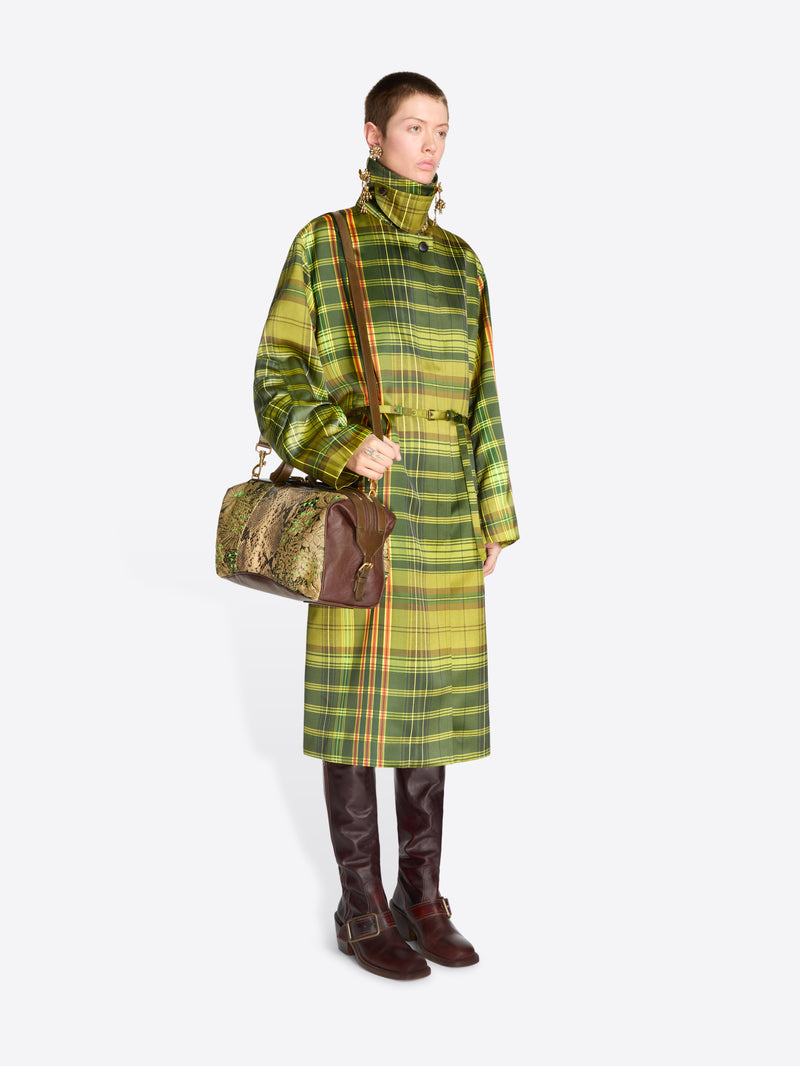 Full-length model wearing a green plaid belted coat, long brass earrings, and dark brown knee-high boots, carrying a floral jacquard duffle bag with tan snakeskin-effect panels.