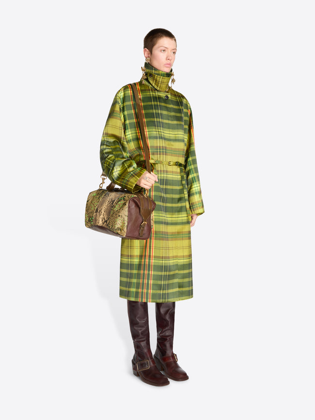 Full-length model wearing a green plaid belted coat, long brass earrings, and dark brown knee-high boots, carrying a floral jacquard duffle bag with tan snakeskin-effect panels.