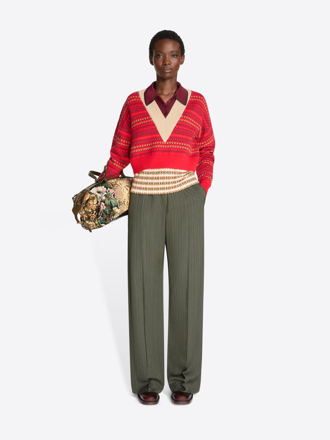 Full-length model wearing a red patterned V-neck sweater and wide-leg olive trousers, carrying a floral jacquard duffle bag with tan snakeskin-effect panels.