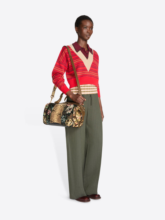 Full-length model wearing a red patterned V-neck sweater and wide-leg olive trousers, holding a floral jacquard duffle bag with tan snakeskin-effect panels.