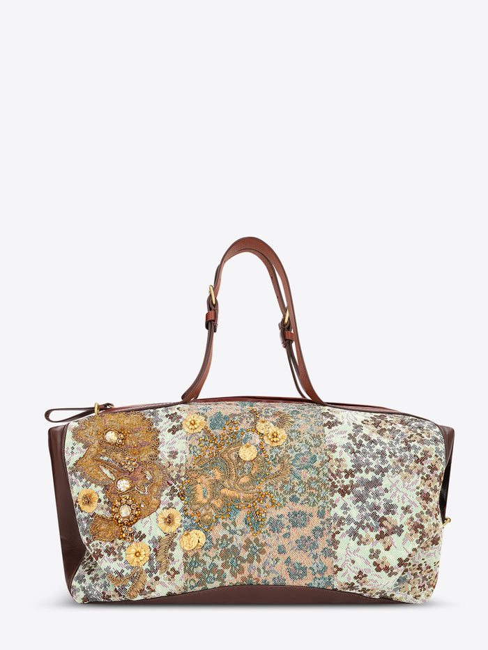 A structured duffel bag with a multicolor floral and paisley patchwork pattern on the front, brown leather handles and side panels, and gold-tone hardware, shown against a plain light gray background.