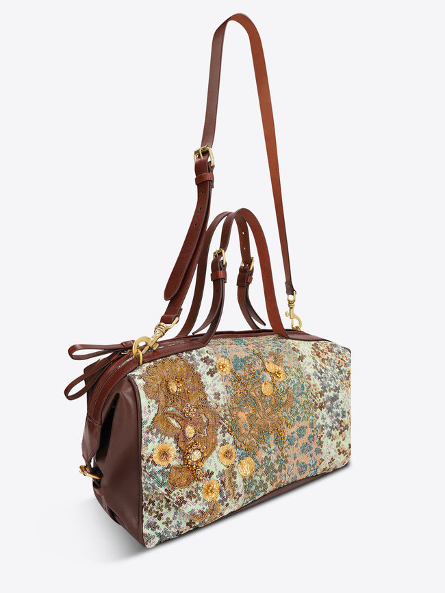 A structured duffel bag with a multicolor floral and paisley patchwork pattern on the front, brown leather handles and side panels, and gold-tone hardware, shown against a plain light gray background.
