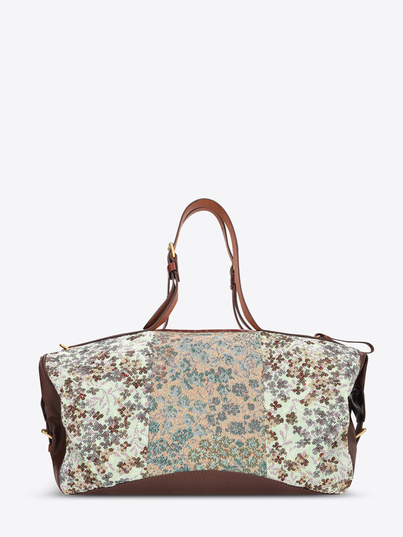 A structured duffel bag with a multicolor floral and paisley patchwork pattern on the front, brown leather handles and side panels, and gold-tone hardware, shown against a plain light gray background.