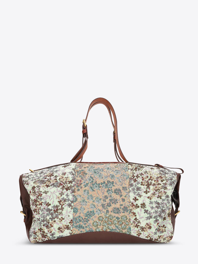 A structured duffel bag with a multicolor floral and paisley patchwork pattern on the front, brown leather handles and side panels, and gold-tone hardware, shown against a plain light gray background.