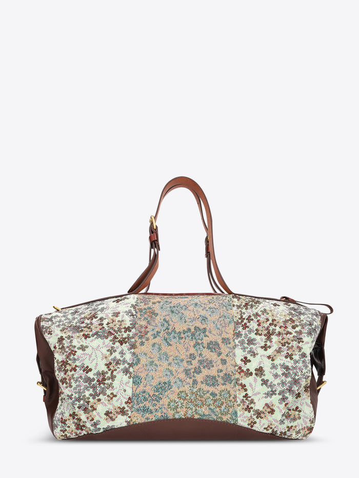 A structured duffel bag with a multicolor floral and paisley patchwork pattern on the front, brown leather handles and side panels, and gold-tone hardware, shown against a plain light gray background.