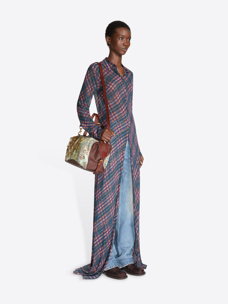 A model stands against a light gray studio background wearing a long-sleeve button-front shirt dress with a red and blue plaid pattern over light blue denim jeans, holding a small brown handbag.