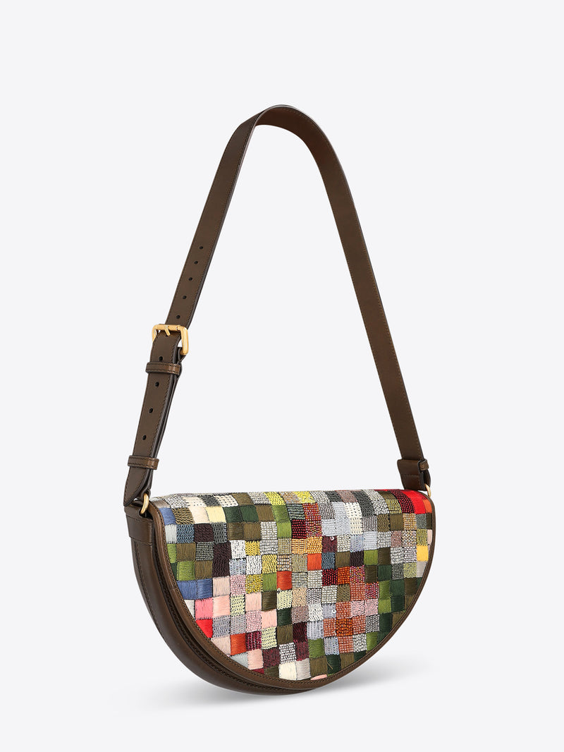 Half-moon shoulder bag with a dark brown leather strap and multicolored textured patchwork panel on a light gray background.