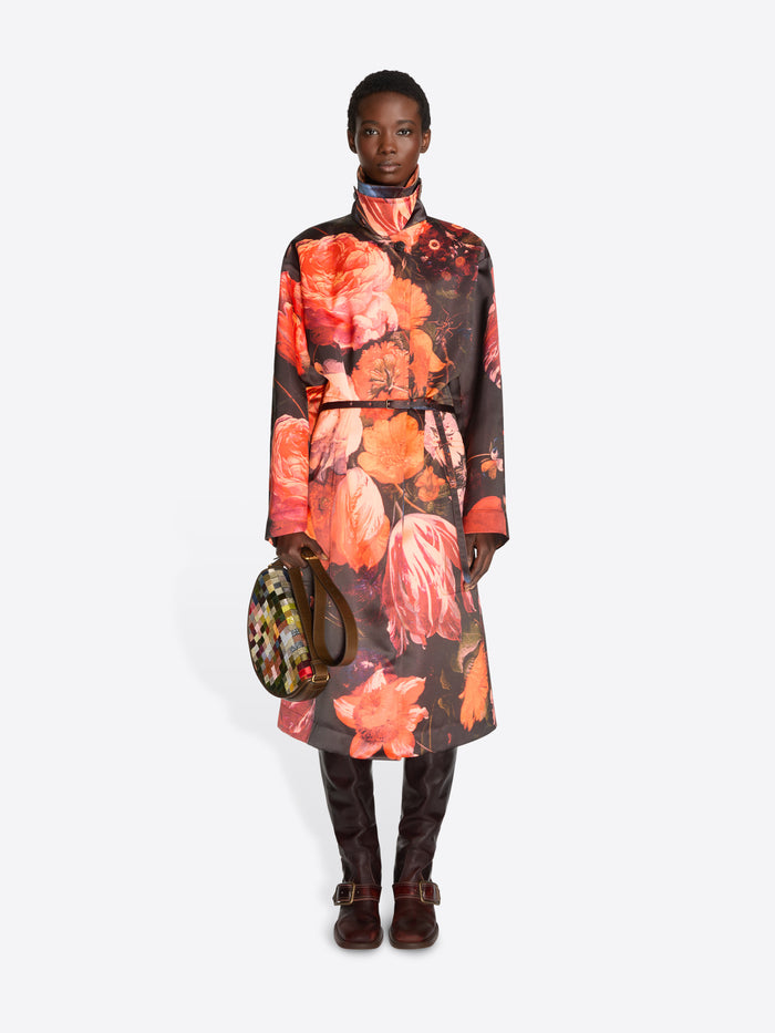 Full-length model wearing a black coat with oversized orange floral print and dark brown knee-high boots while carrying a multicolored checkered half-moon bag.