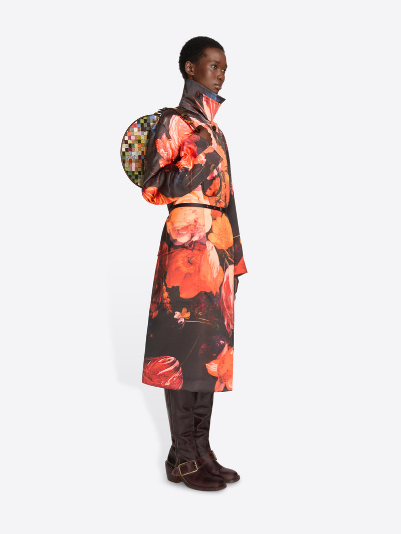 Full-length model wearing a black coat with oversized orange floral print and dark brown knee-high boots while carrying a multicolored checkered half-moon bag.