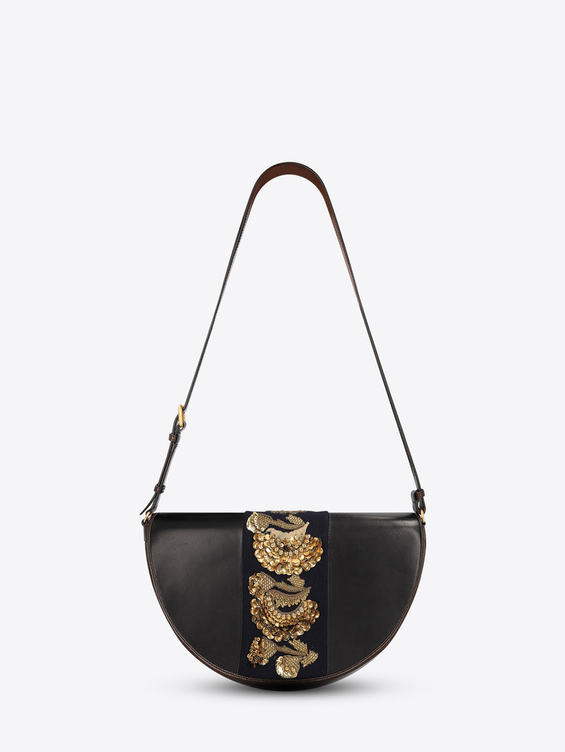 Black leather half-moon shoulder bag with a dark brown strap and a navy center panel featuring ornate gold-toned embroidery and sequins on a light gray background.