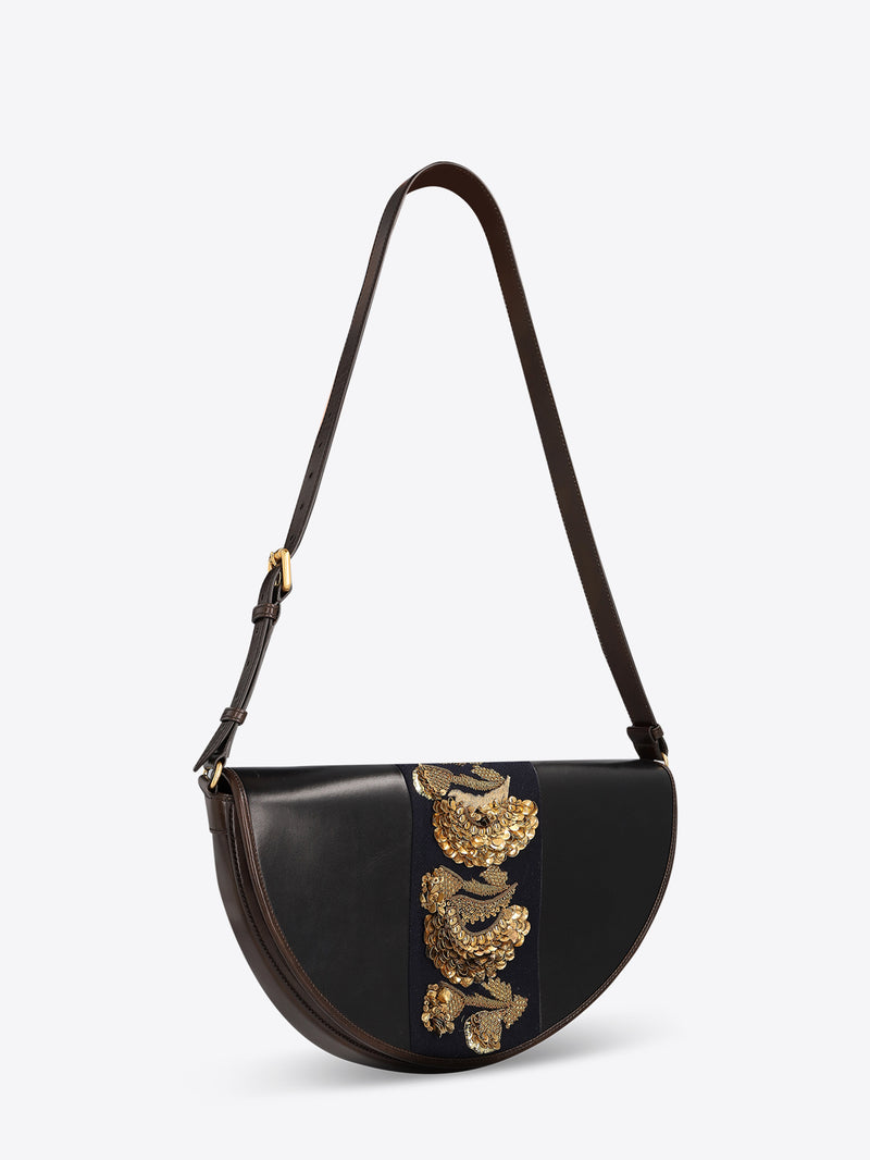 Black leather half-moon shoulder bag with a dark brown strap and a navy center panel featuring ornate gold-toned embroidery and sequins on a light gray background.