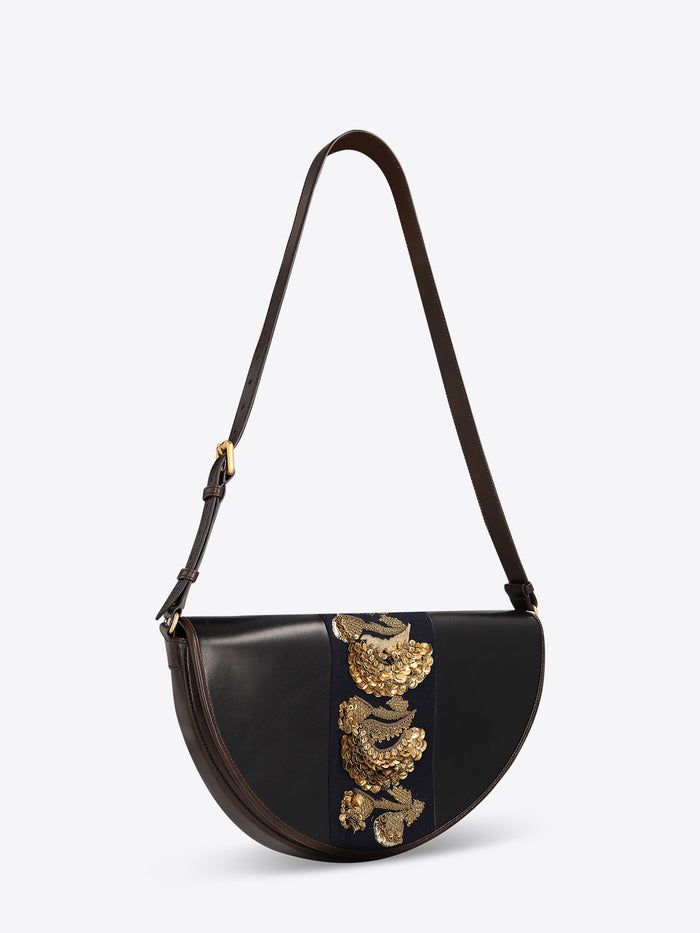 Black leather half-moon shoulder bag with a dark brown strap and a navy center panel featuring ornate gold-toned embroidery and sequins on a light gray background.