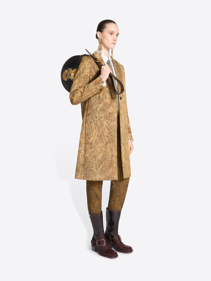 Full-length model wearing a gold paisley coat with matching trousers, a white shirt and tie, long brass earrings, and dark brown boots while carrying a black half-moon bag with ornate gold embroidery.