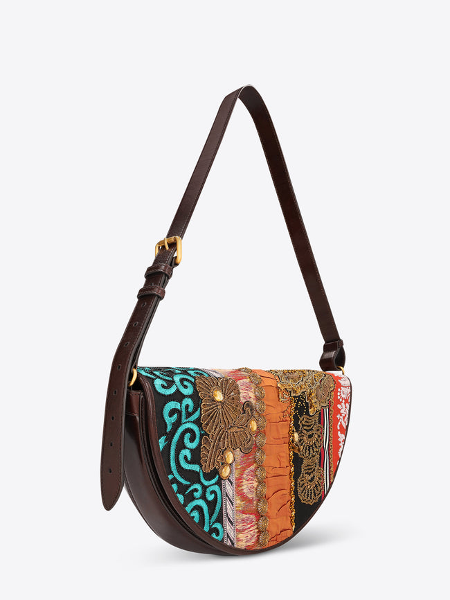 Half-moon shoulder bag with a dark brown leather strap and multicolored patchwork panels featuring embroidery, beading, and floral appliqués on a light gray background.