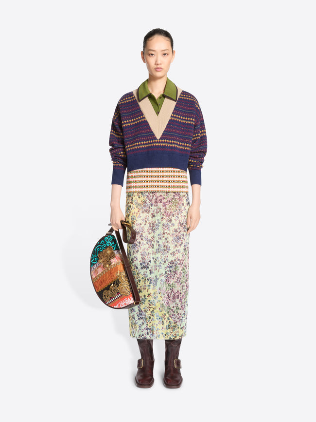 Full-length model wearing a patterned V-neck sweater, floral midi skirt, and brown boots while carrying a multicolored patchwork half-moon shoulder bag.