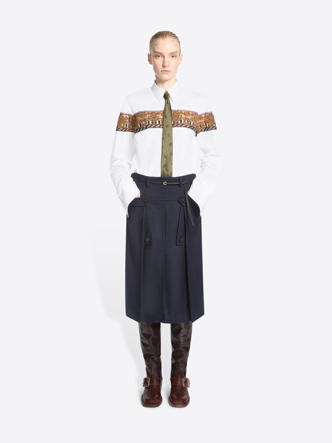 A model stands against a light gray background wearing a white long-sleeve shirt with a wide brown patterned band across the chest, an olive patterned tie, a dark navy high-waisted belted skirt, and brown knee-high boots.