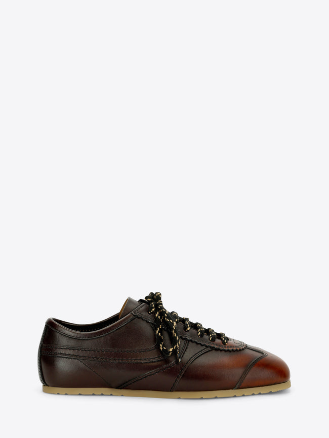 A dark brown leather low-top sneaker with decorative laces, a brown leather toe cap, and a light tan rubber sole, shown against a light gray background.