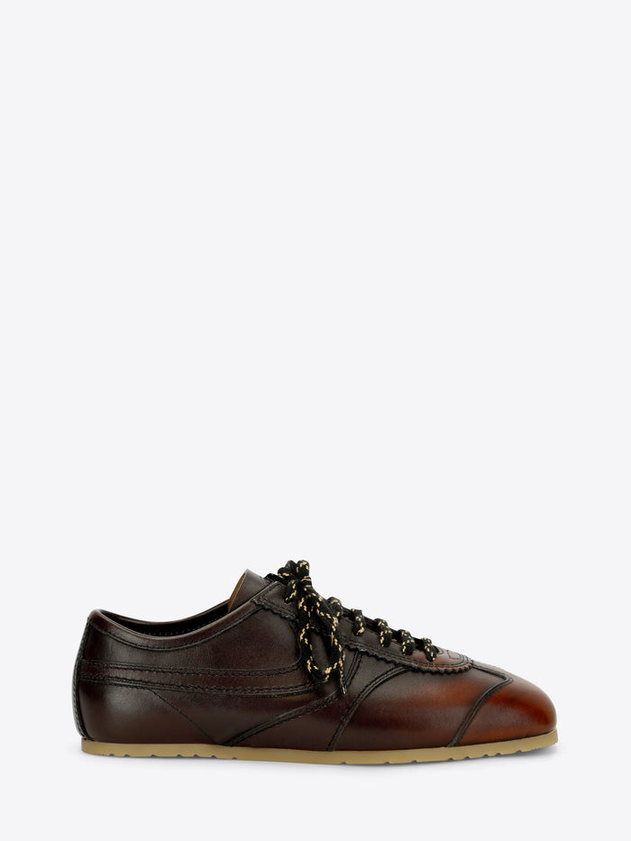 A dark brown leather low-top sneaker with decorative laces, a brown leather toe cap, and a light tan rubber sole, shown against a light gray background.