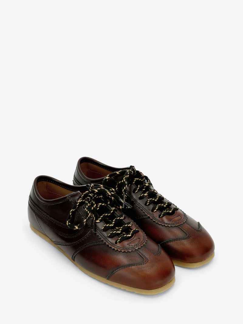 A pair of dark brown leather low-top sneakers with decorative laces, a brown leather toe cap, and a light tan rubber sole, shown against a light gray background.
